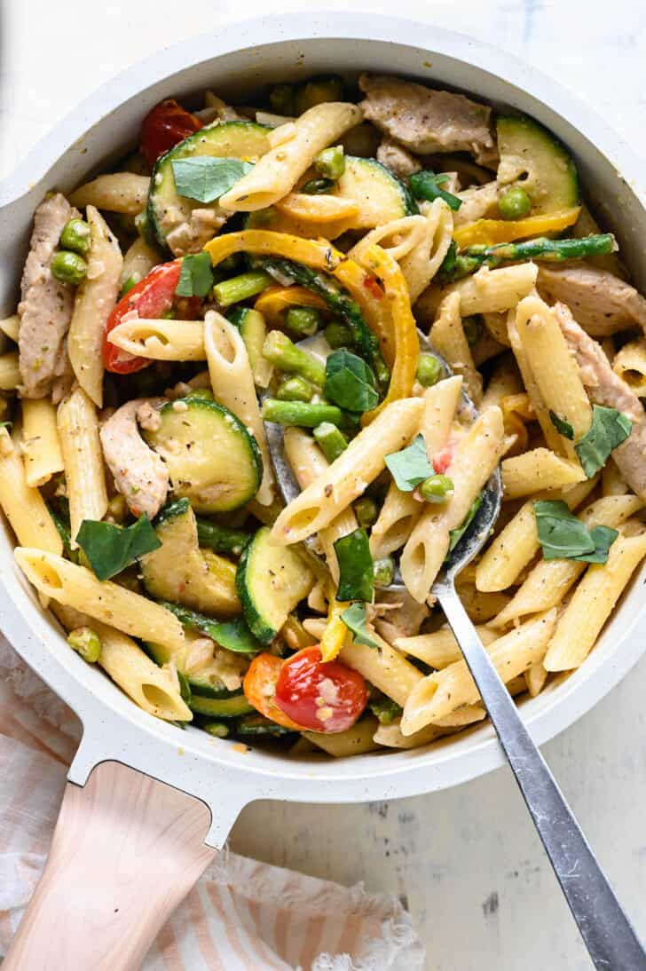 A white pot filled with pasta primavera with chicken, with a spoon digging into it.