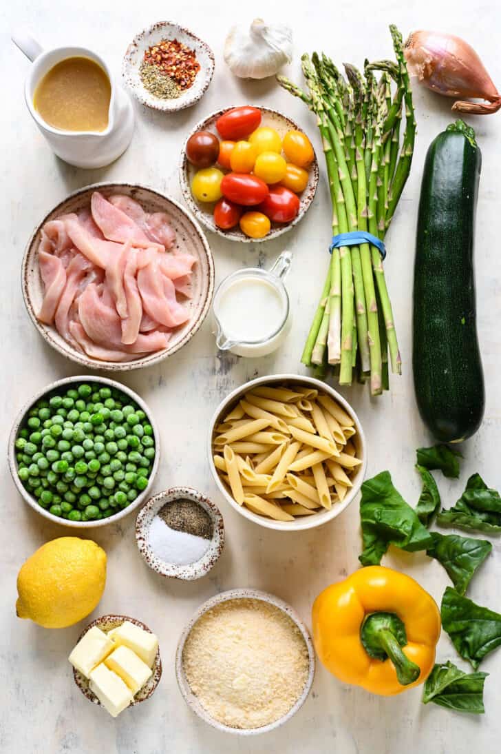 Ingredients laid out on a light surface including chicken, spices, cream, Parmesan cheese, butter, frozen peas, penne pasta, and lots of fresh vegetables.