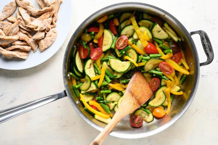 A stainless steel skillet filled with sliced zucchini, asparagus, grape tomatoes and yellow pepper, with a wooden spoon stirring it.