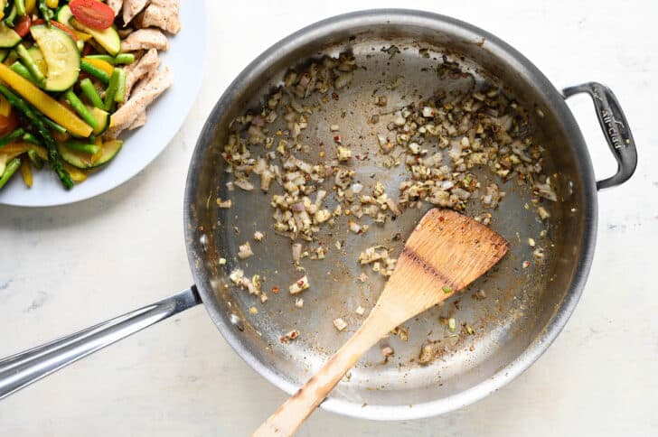 A stainless steel skillet filled with minced shallots and garlic and dried spices, with a wooden spoon stirring it.