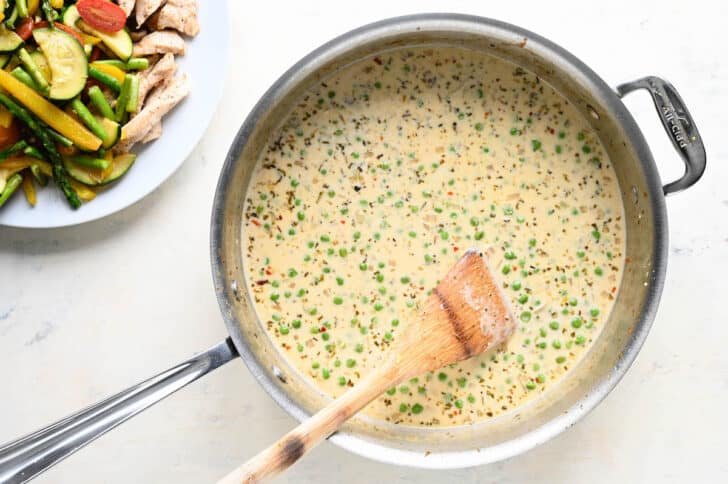 A stainless steel skillet filled with a creamy sauce with green peas in it, with a wooden spoon stirring it.