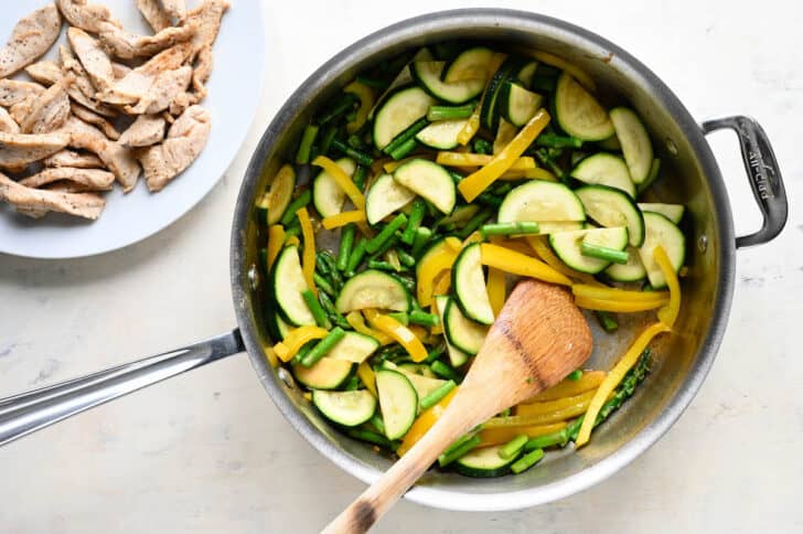 A stainless steel skillet filled with sliced zucchini, asparagus and yellow pepper, with a wooden spoon stirring it.