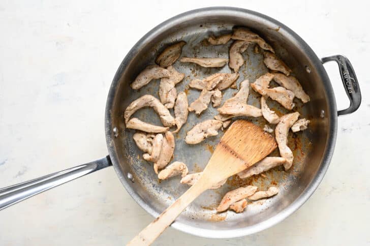 A stainless steel skillet filled with sauteed slices of chicken, with a wooden spoon stirring it.