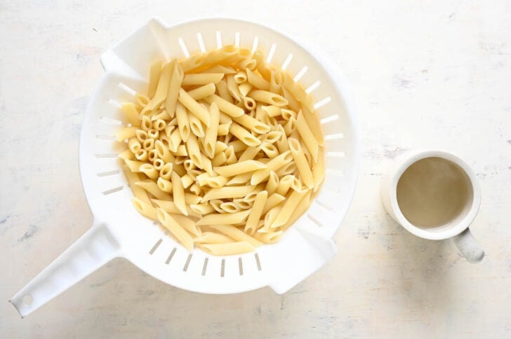 A white colander filled with cooked penne pasta, alongside a steaming mug of water.