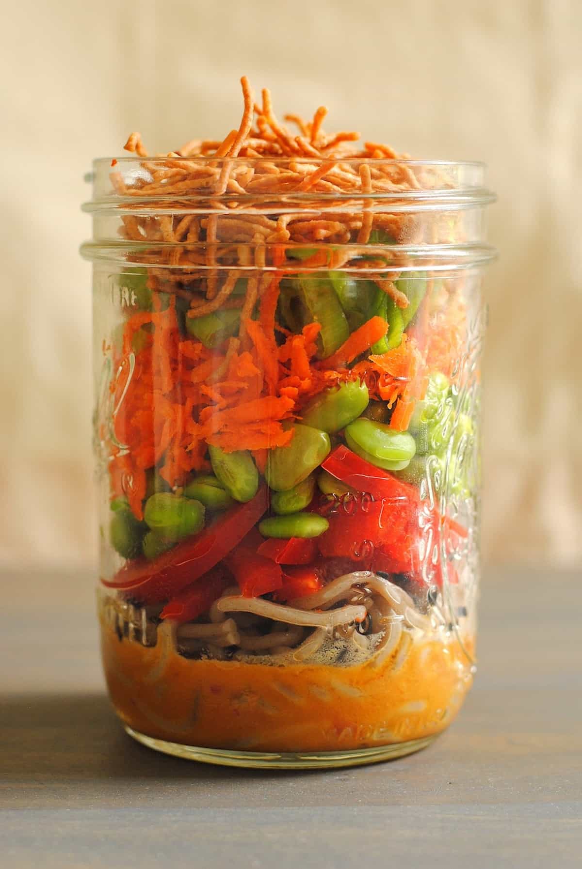 Glass mason jar filled with layers of dressing, noodles and vegetables.
