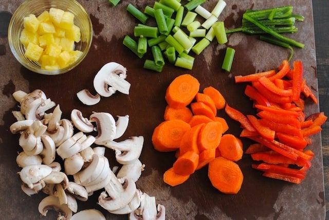 Brown cutting board with sliced mushrooms, carrots, red pepper, jalapenos, green onions and pineapple.