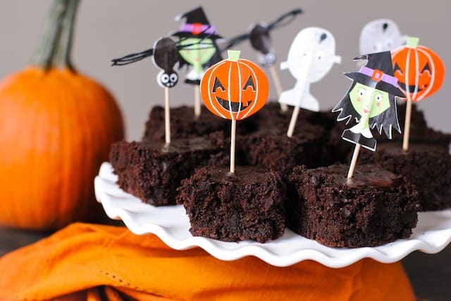Brownies on white platter with orange napkin and pumpkin decorating scene. Pumpkin, witch, bat and ghost decorative toothpicks are stuck in brownies.