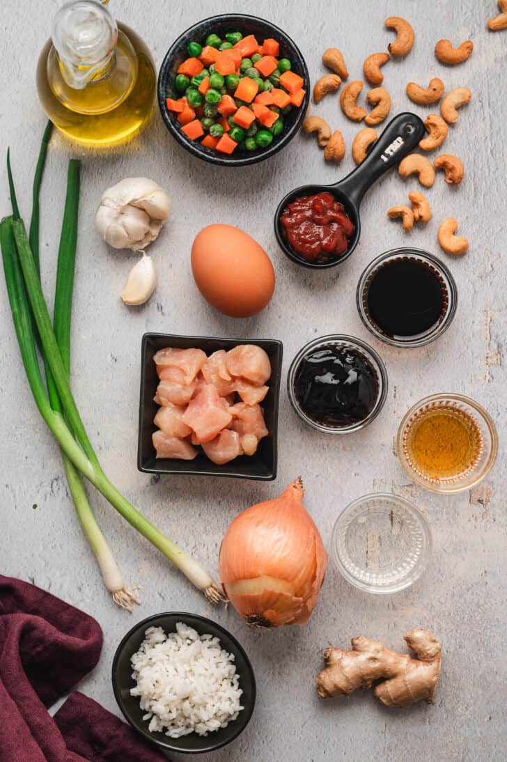 Ingredients laid out on a light surface including green onions, garlic, peas and carrots, chicken, rice, an egg, cashews and Asian condiment sauces.
