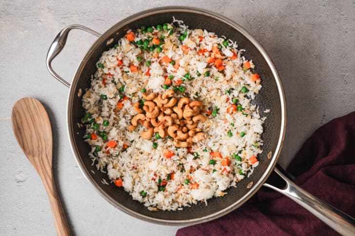 A dark nonstick skillet filled with rice and chopped vegetables, and cashews.