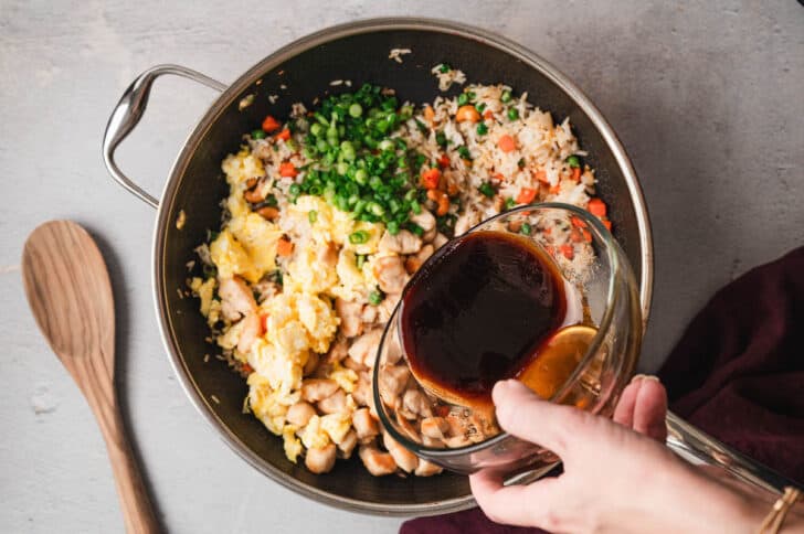 A dark nonstick skillet filled with an easy chicken fried rice recipe, with a hand pouring dark sauce into the skillet.