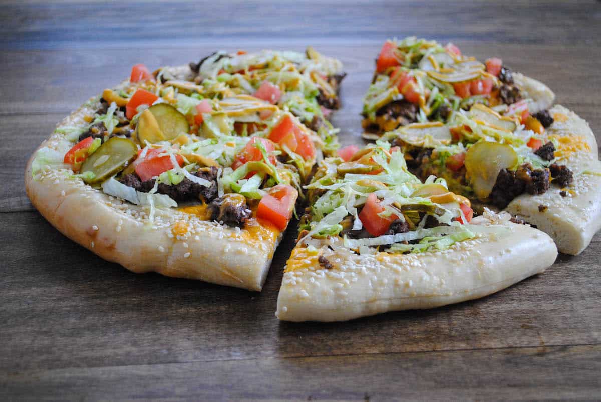 Horizontal photo of round flatbread topped with burger toppings, cut into six slices.