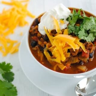 Chili topped with shredded cheddar cheese, sour cream and cilantro in white bowl, on top of white plate with spoon. Cheese and cilantro scattered on table near bowl.