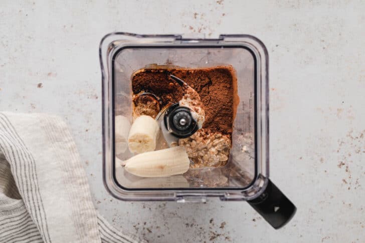 A square blender pitcher filled with oats, cocoa and banana.