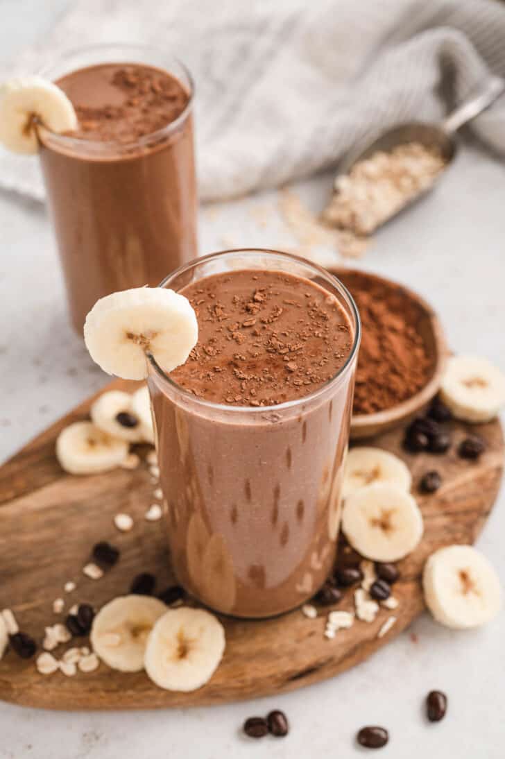 Two glasses of a coffee breakfast smoothie recipe, garnished with cocoa powder and sliced bananas.