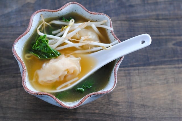 A small rustic bowl is filled with broth, dumplings and greens, with a white soup spoon scooping one dumpling.