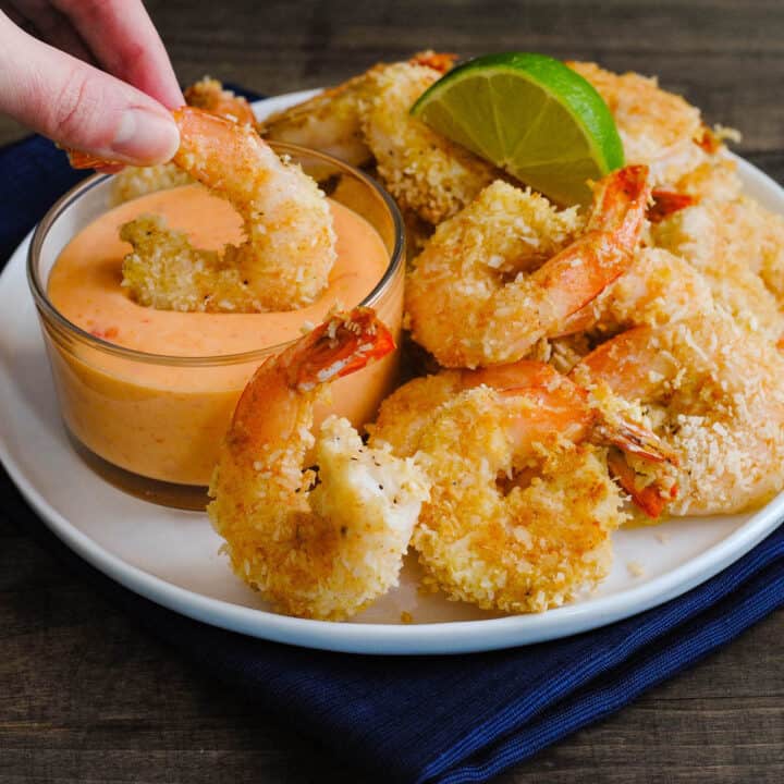 A white plate topped with coconut shrimp, with a hand dipping one shrimp into a glass ramekin of coconut shrimp sauce.