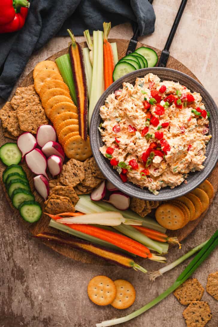 A round wooden cutting board with a bowl of pimento cheese dip surrounded by crackers and cut vegetables.