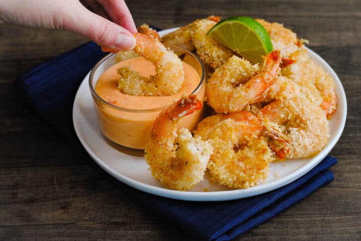 A white plate topped with fried prawns, with a hand dipping one prawn into a glass ramekin filled with a creamy orange mixture.