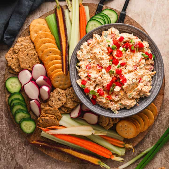 A round wooden cutting board with a bowl of pimento cheese dip surrounded by crackers and cut vegetables.