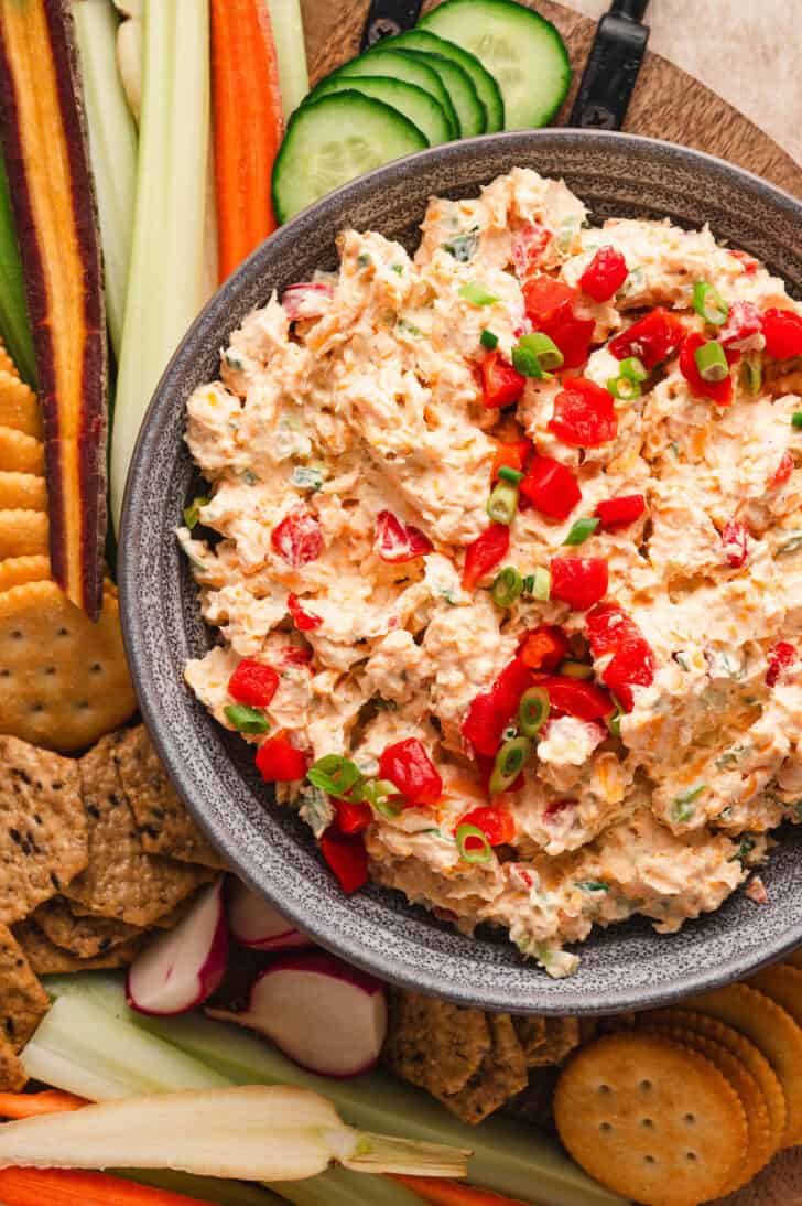 Pimento cheese spread in a gray bowl surrounded by crackers and cut vegetables.