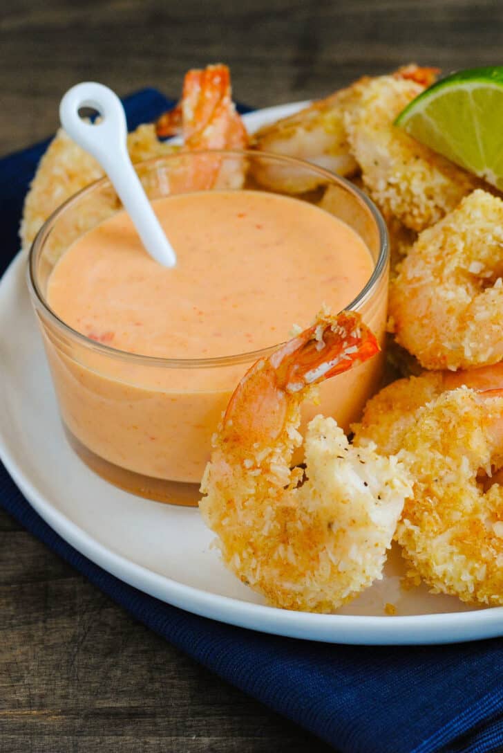 A white plate topped with coconut shrimp and  glass ramekin of sauce for coconut shrimp.