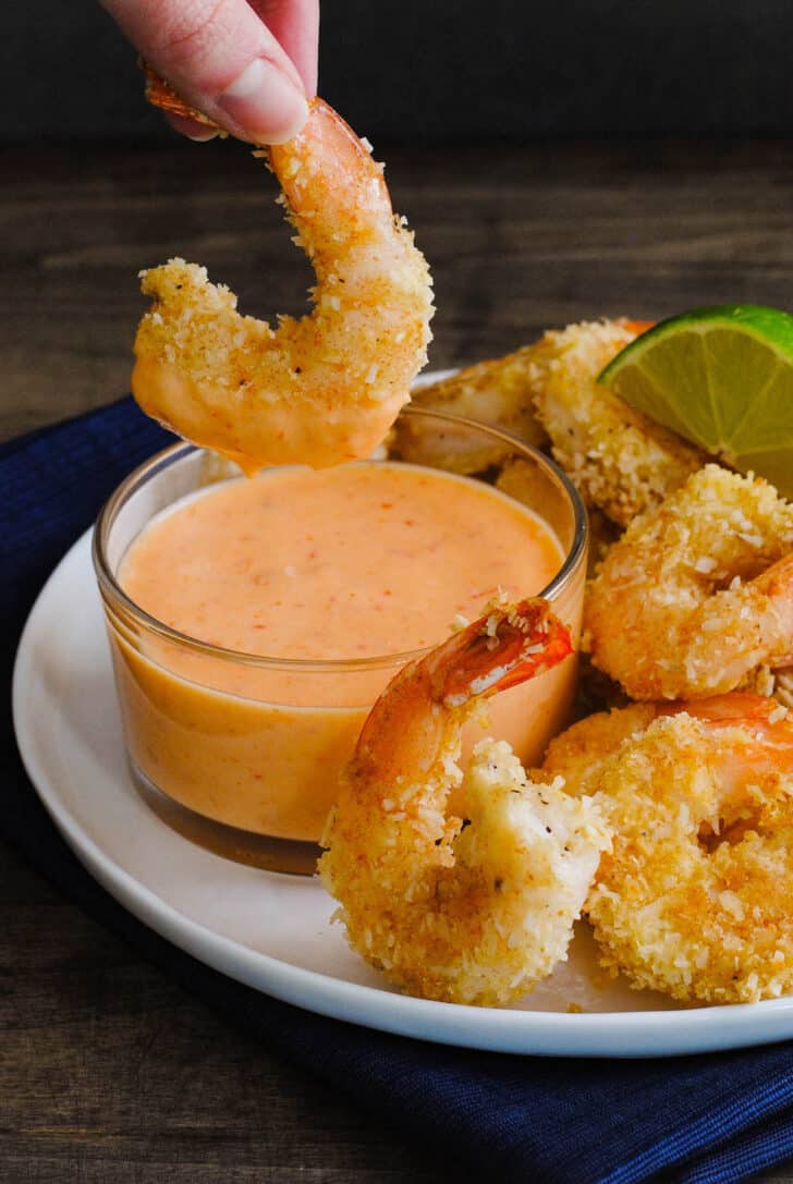 A white plate topped with coconut shrimp, with a hand dipping one shrimp into a glass ramekin of coconut shrimp sauce.