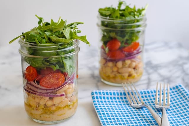 Two mason jars layered with marinated cannelini beans, sliced red onion, grape tomatoes and arugula. On light marble countertop with blue napkin and fork.