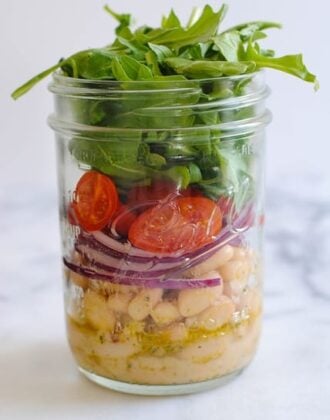 Mason jar layered with marinated white beans, sliced red onion, grape tomatoes and arugula. On light marble countertop.