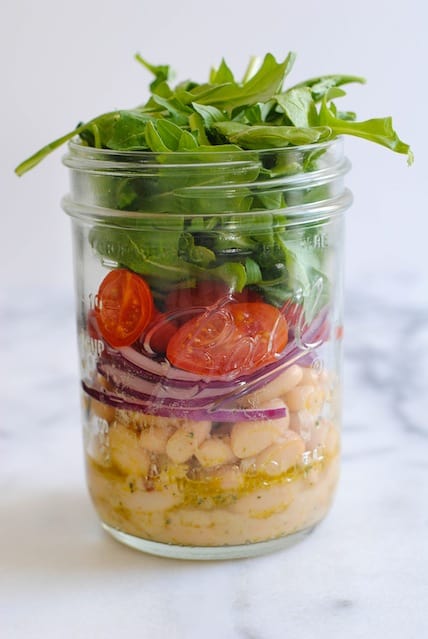 Mason jar layered with white beans salad, sliced red onion, grape tomatoes and arugula. On light marble countertop with blue napkin and fork.