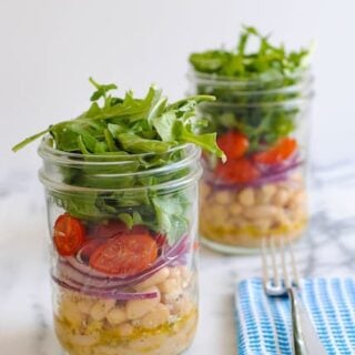 Two mason jars layered with marinated white beans, sliced red onion, grape tomatoes and arugula. On light marble countertop with blue napkin and fork.