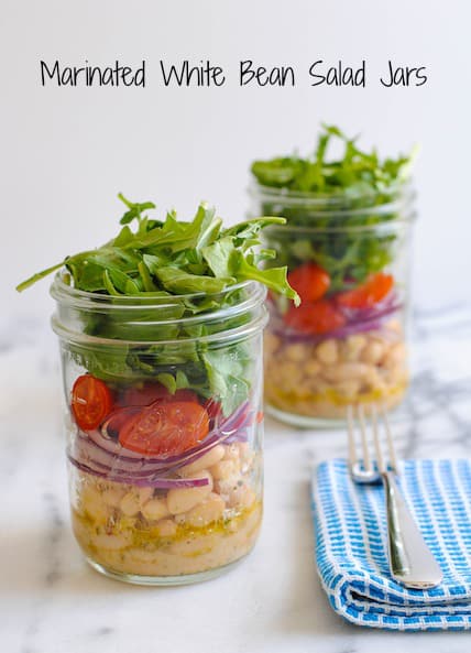 Two mason jars layered with marinated white beans, sliced red onion, grape tomatoes and arugula. On light marble countertop with blue napkin and fork.