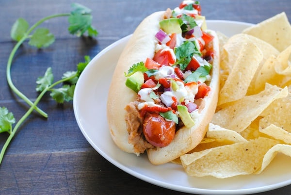 A hot dog with Mexican condiments on a plate with tortilla chips.