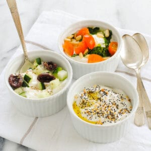 Three white ramekins of savory Greek yogurt recipes, including bowls topped with everything bagel seasoning, olives and cucumbers, and tomato, basil pesto and pine nuts.