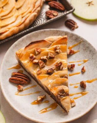 A slice of apple tart drizzled with caramel and sprinkled with chopped pecans, on a small plate.