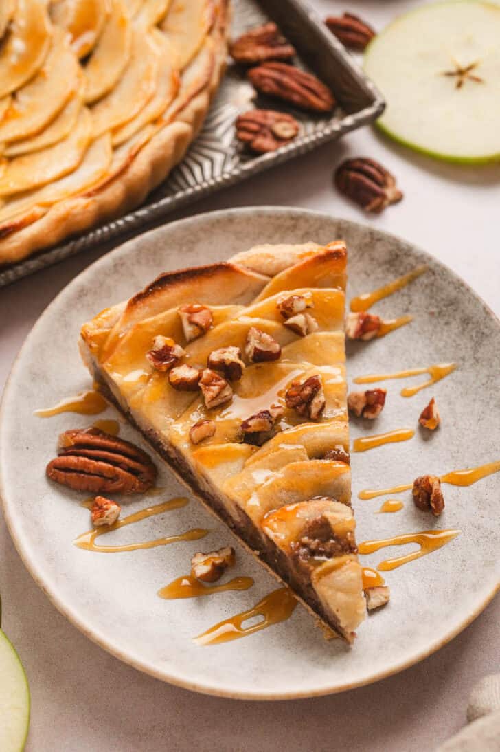 A slice of apple tart drizzled with caramel and sprinkled with chopped pecans, on a small plate.