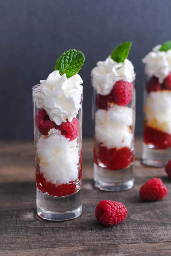 Closeup on shooter glass filled with layers of angel food cake, jam, raspberries, whipped cream, topped with mint leaf.