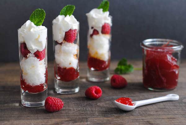 Three shooter glasses filled with sweet items like cake, berries and whipped cream, alongside more fresh berries and jar of jam.