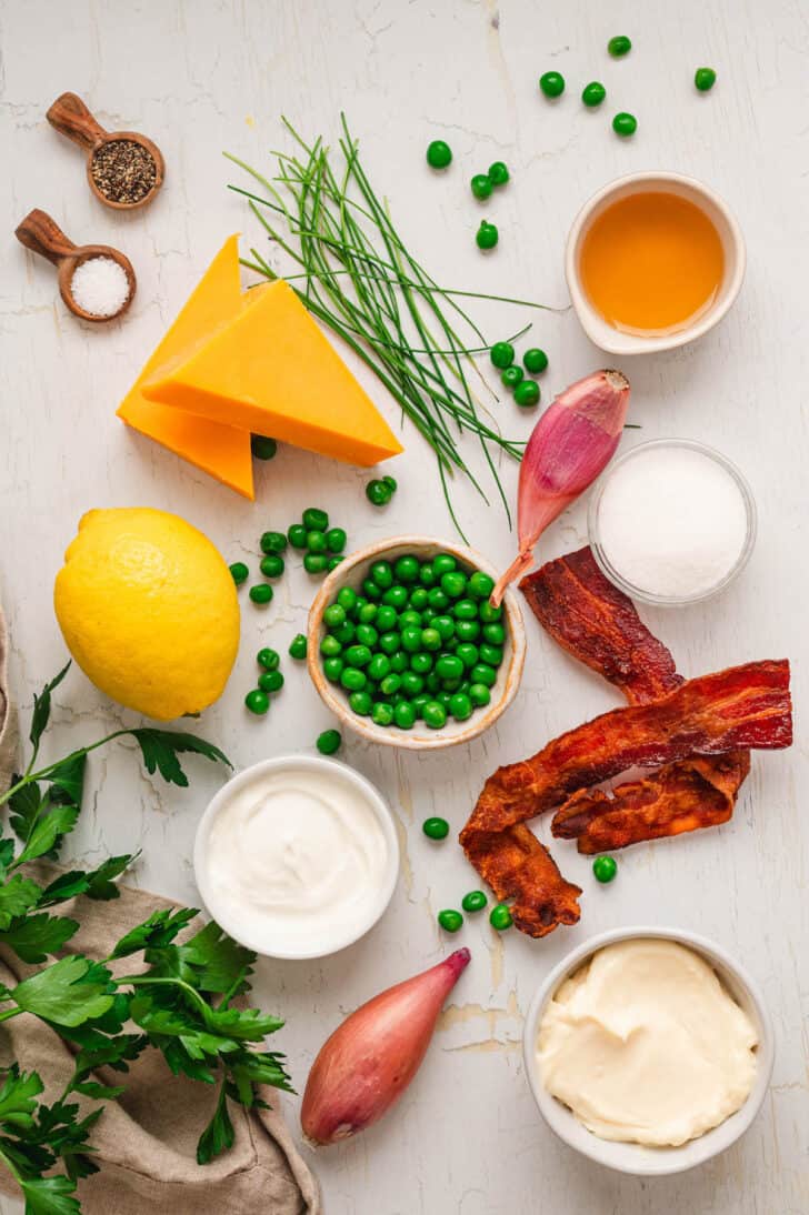Ingredients laid out on a light surface, including peas, bacon, shallots, herbs, condiments, spices and cheese.