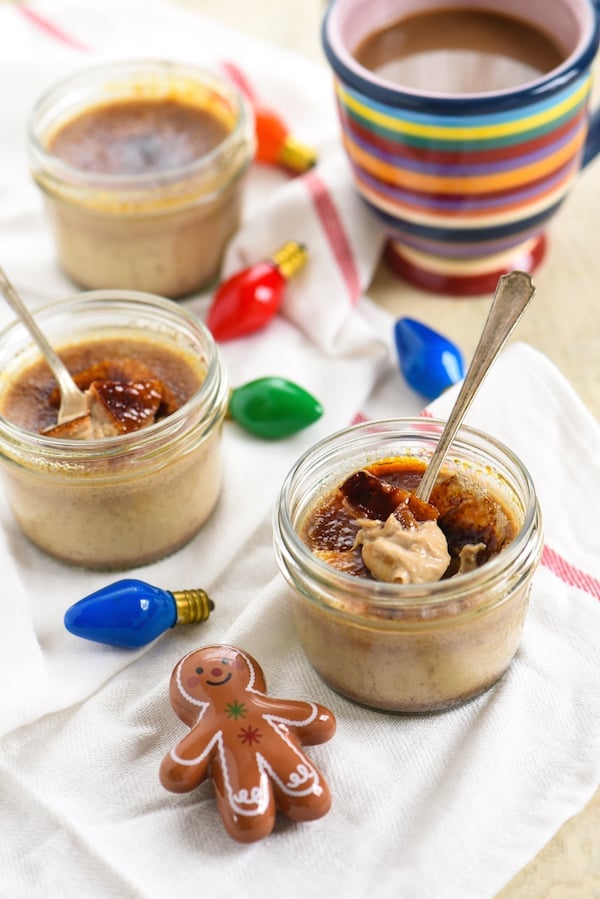 Small mason jars filled with torched ginger bread custard, with spoons in them. Christmas tree lightbulbs and a ceramic gingerbread person decorate the scene.