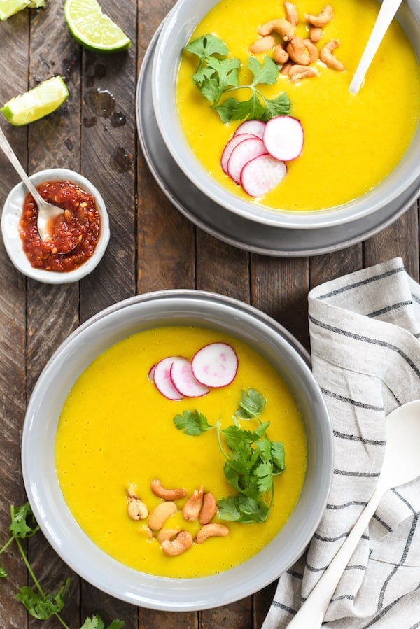 Overhead photo of two gray bowls filled with bright orange soup. Soups are topped with cashews, cilantro and radishes, and there is a small bowl of hot sauce on the table.