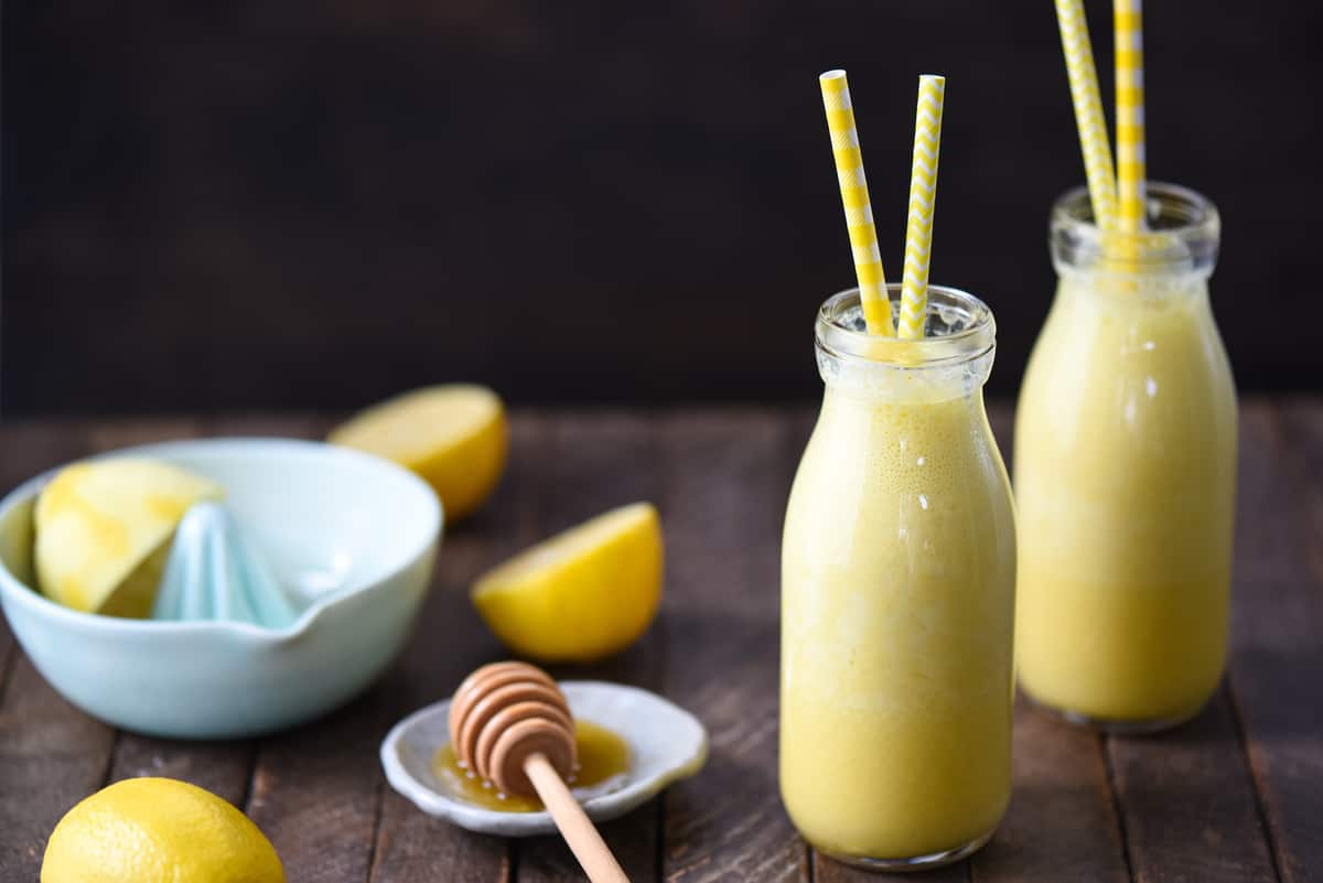 Two small glass bottles filled with lemon ginger smoothie, with yellow straws sticking out of the bottles. A citrus juicer and a honey dish are in the background.