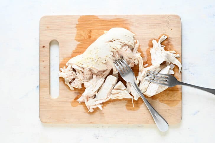 A wooden cutting board with a cooked chicken breast on it, with two forks shredding it.