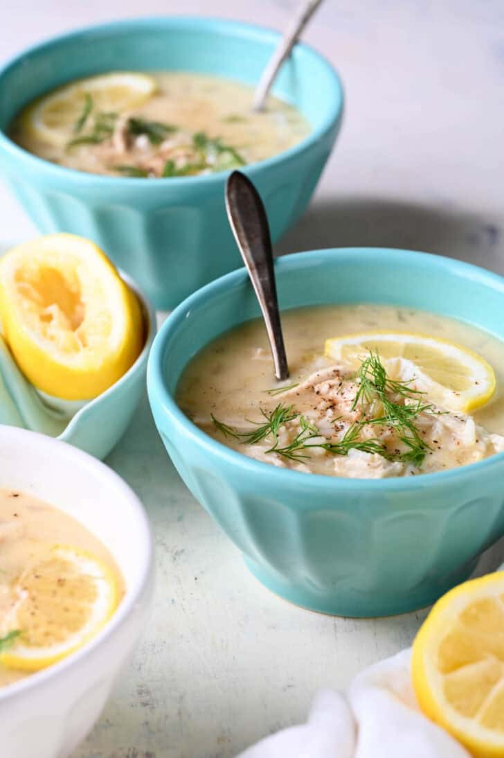 Teal bowls filled with an avgolemono soup recipe garnished with lemon wedges and fresh dill.