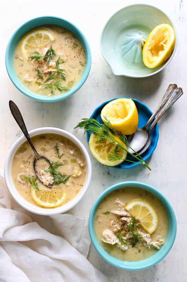 A light surface filled with bowls of Greek lemon soup, with fresh lemons and dill garnishing the scene.