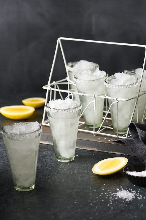 Set of small, tall glasses, filled with ice and a clear beverage, with cut lemons and salt nearby.