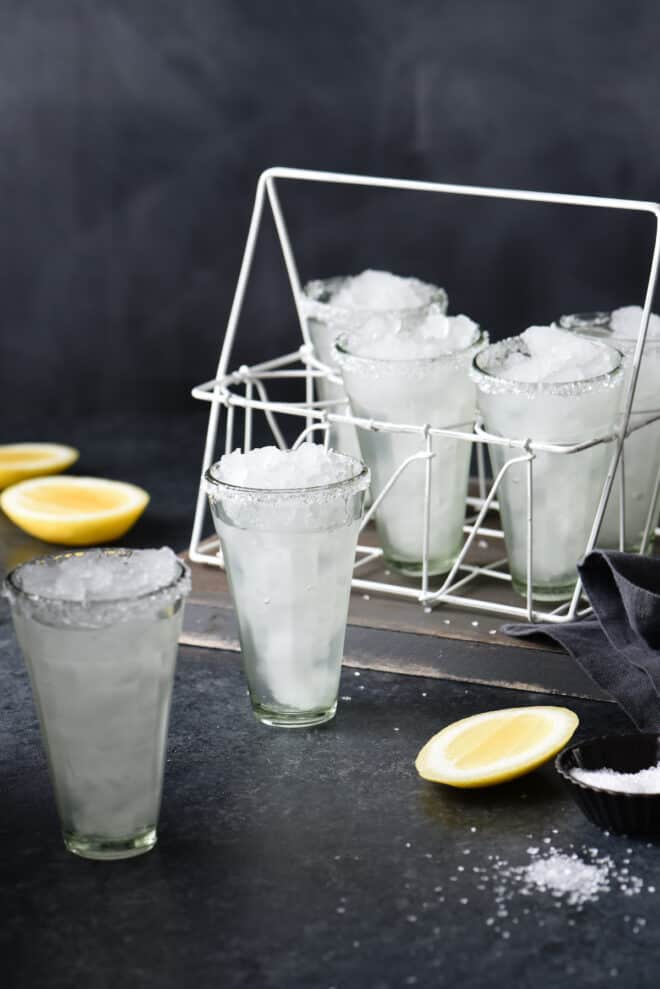 Set of small, tall glasses, filled with ice and a clear beverage, with cut lemons and salt nearby.