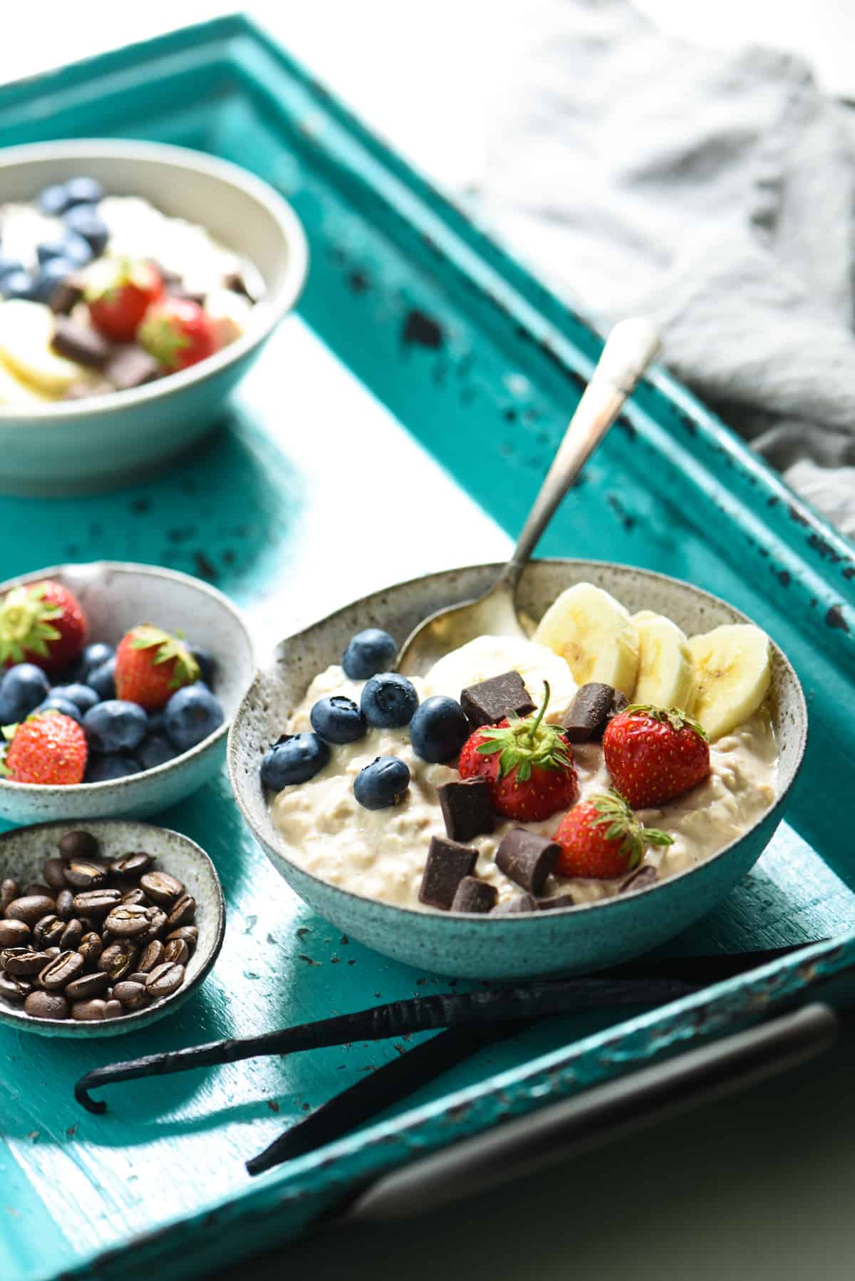 A bowl of overnight oats with yogurt, topped with bananas, blueberries and sliced banana, on a rustic teal tray.
