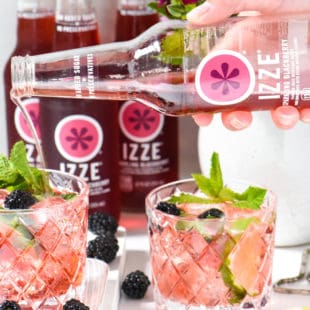 Two small glasses of a pink cocktail garnished with mint and blackberries, with a hand pouring blackberry soda into one of the glasses.