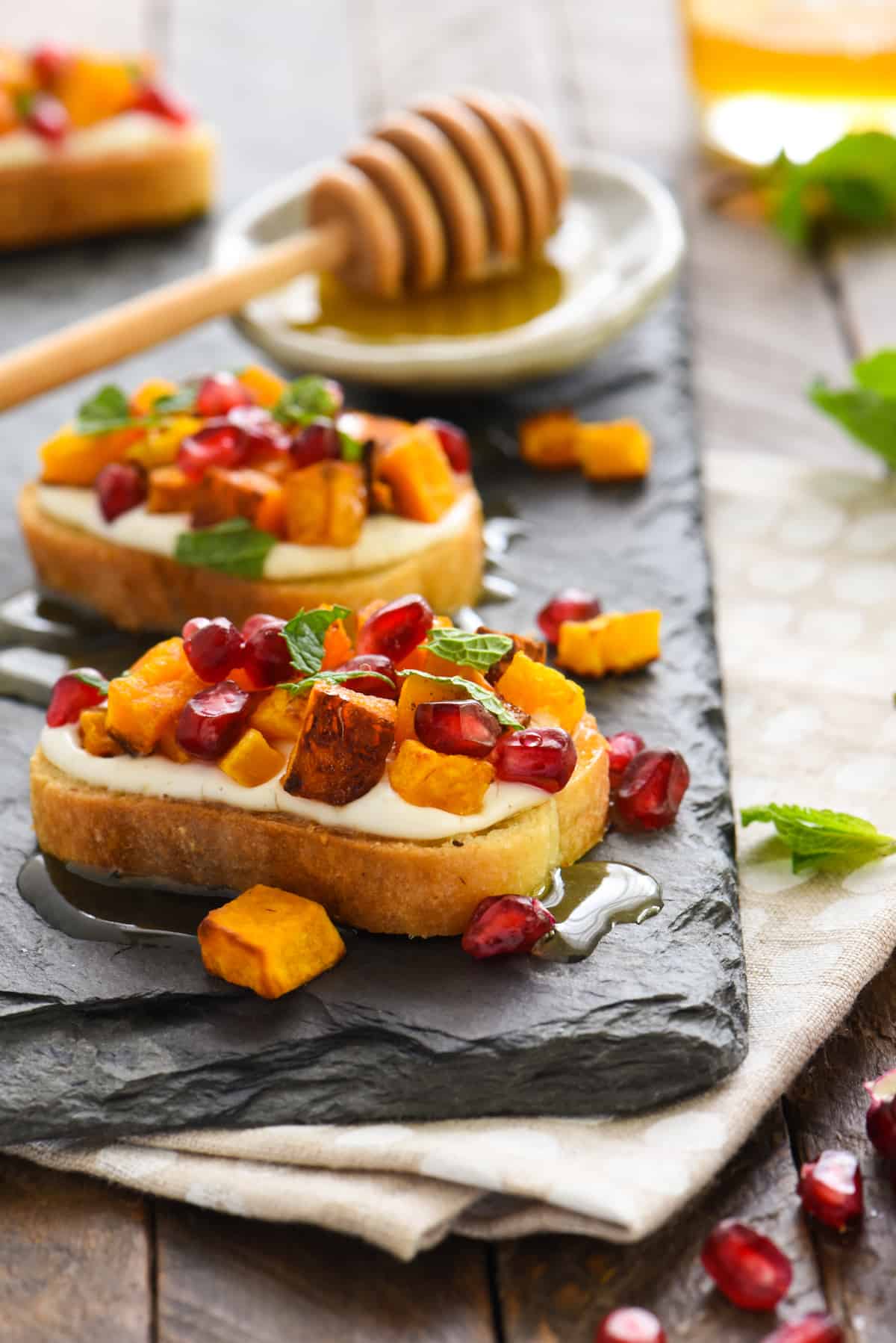 Crostini topped with white sauce, butternut squash and pomegranate seeds, on slate platter.