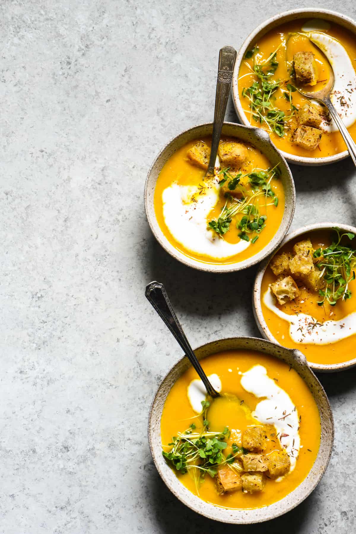 Overhead shot of 4 bowls of soup made with apples and carrots on gray background, topped with yogurt, herbs and croutons.